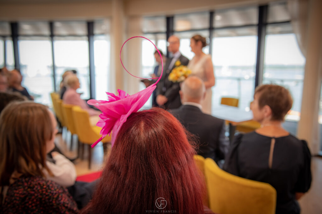 Woman with red hair wearing a pink wedding hat looking towards couple in a bright room