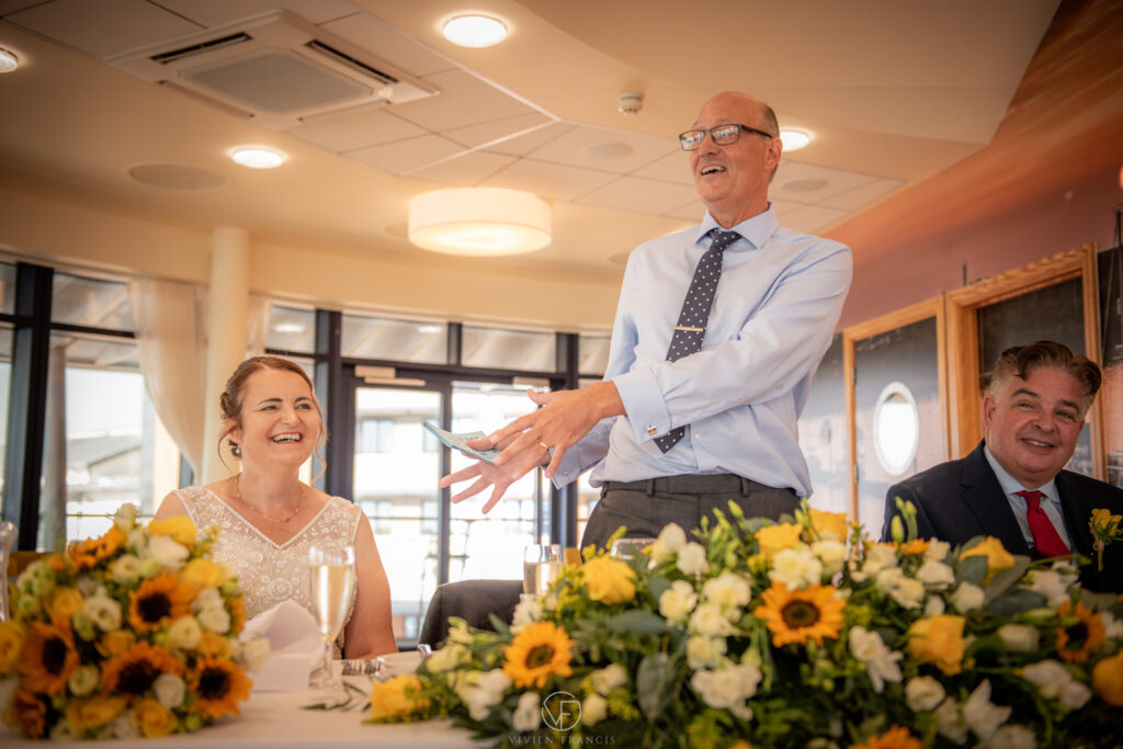 Groom standing up speaking and making the bride smile at a table full of flowers