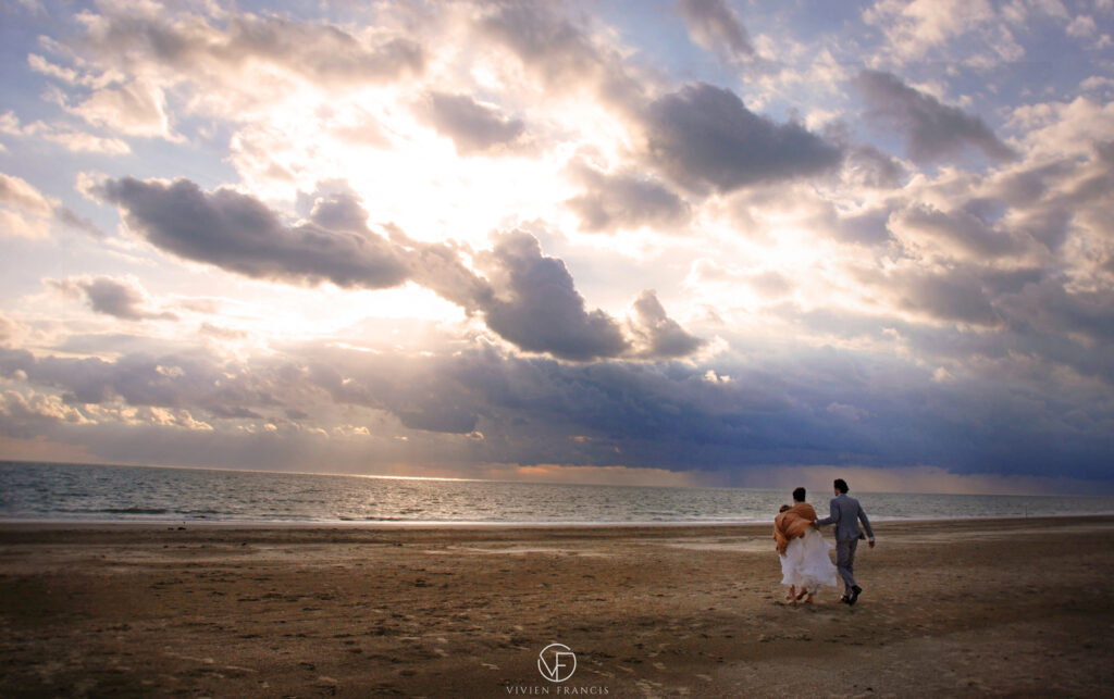 Woman in white dress with child and man in suit walking on the beach towards the sea looking at a sunset and moody clouds