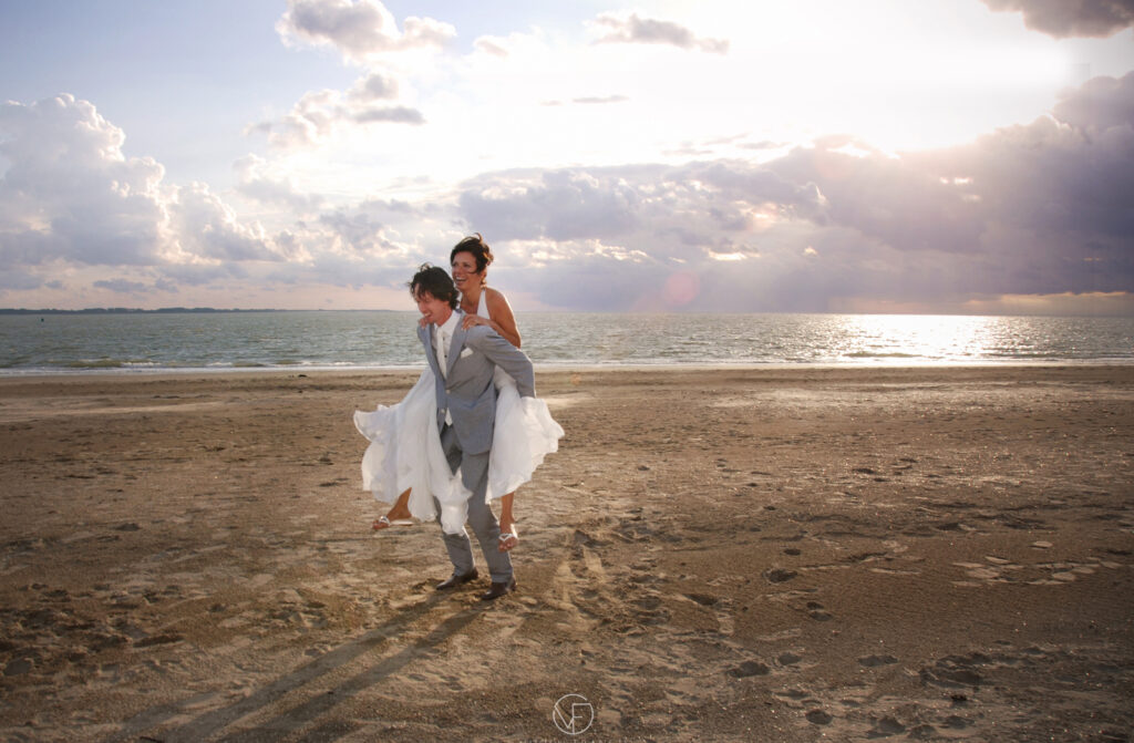 Groom holding the bride on his back laughing together on a beach at sunset
