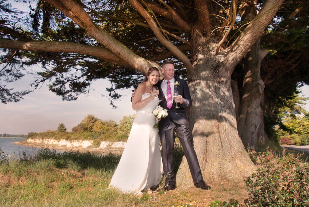 Bride and groom standing together by a tree holding champagne glasses and smiling
