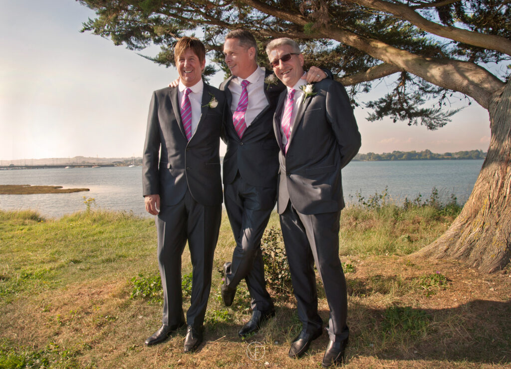 Three men in identical suits with flowers in pockets posing and smiling with a tree in the background