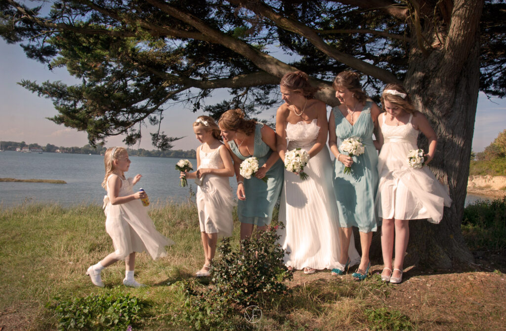 Little girl in white dress joins the other bridesmaids and bride standing outside by a tree
