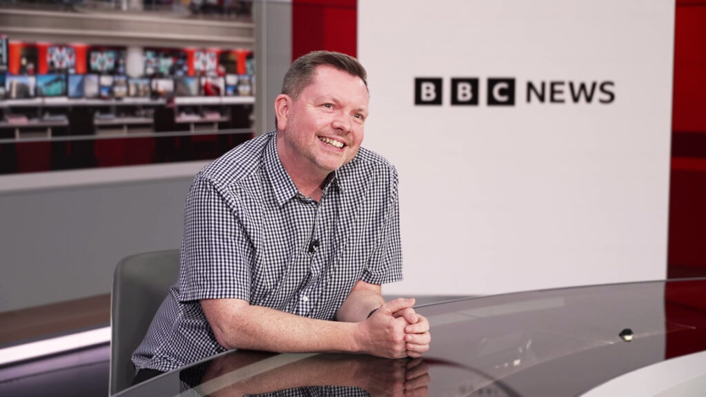 A man sitting in a newsroom, smiling and with the words BBC News in the background