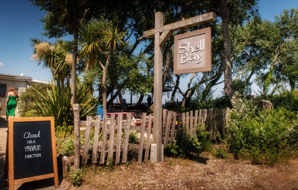 Outside restaurant wooden sign saying Shell Bay with palm trees, wooden fence and sea view