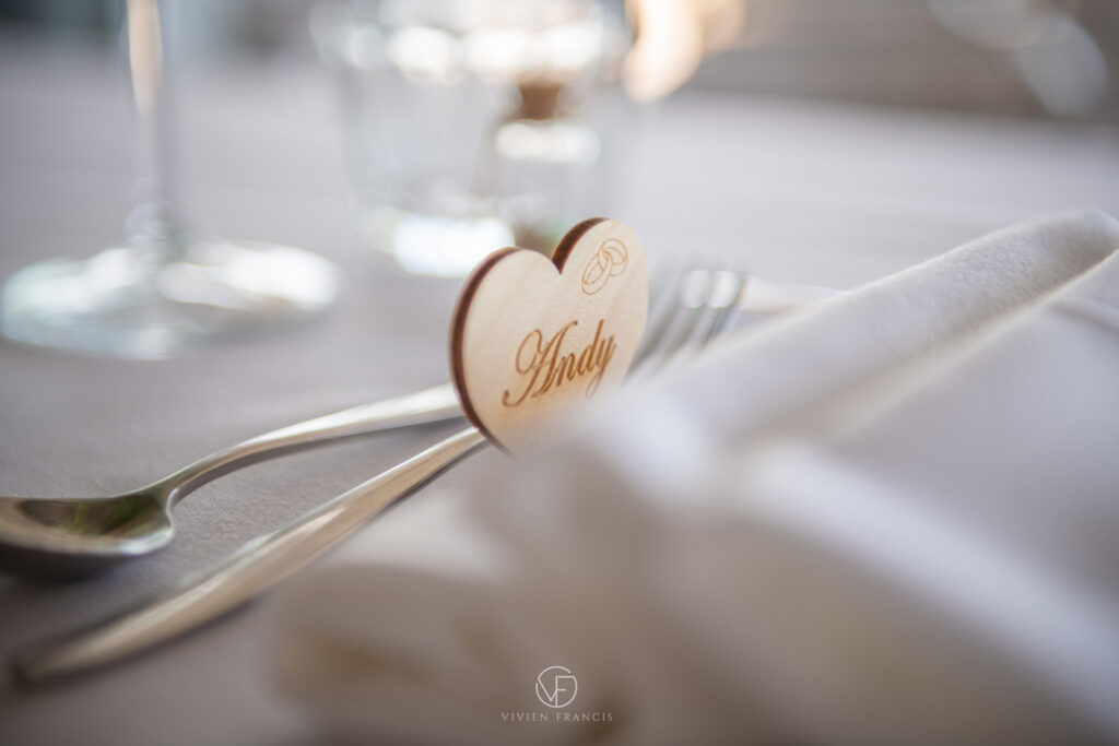 Wooden heart-shaped name tag on a white linen table, with cutlery and glass in the background