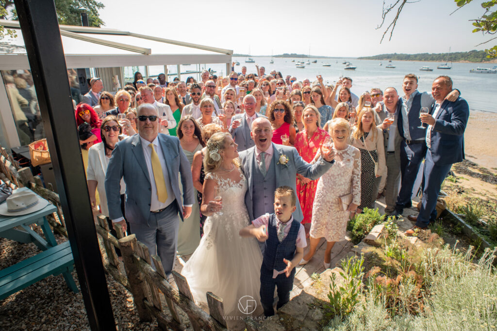 Large group of wedding guests standing outside raising a glass with the sea and boats in the background