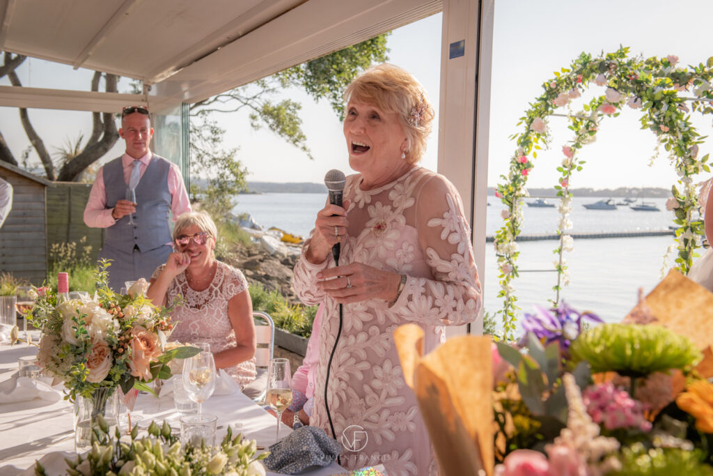 Mother of bride in white soft pink dress holding a microphone standing by a dinner table
