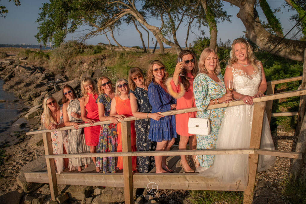 Group of female wedding guests standing in a line behind bride on wooden stairs, with trees and harbour in the background