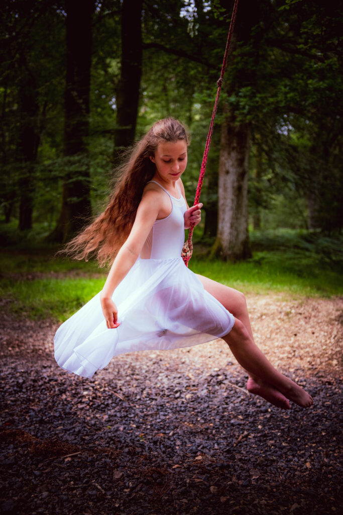 Young girl holding her white dress on a swing rope in a forest with her hair in the wind