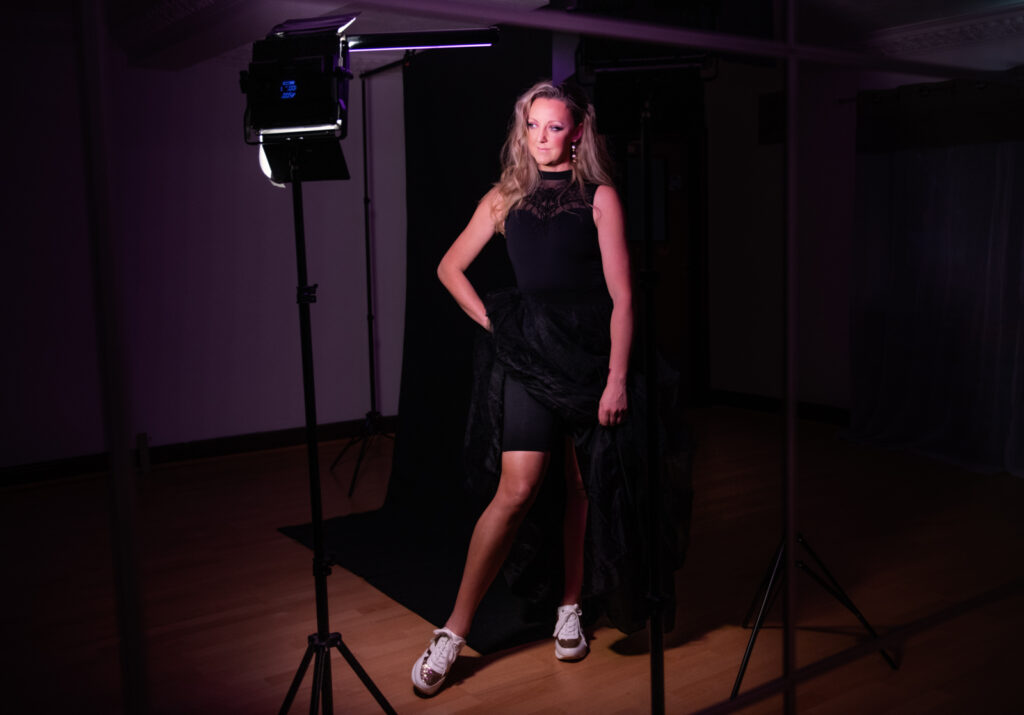 Lady looking into a light with black dress and white sporty trainers with gold jewellery and dark purple eye shadow